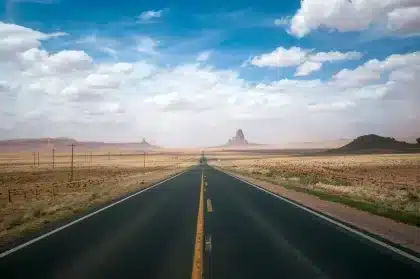 Schnurgerade Wüstenstraße unter weitem Himmel mit roten Felsen am Horizont