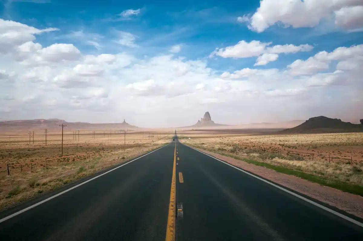 Schnurgerade Wüstenstraße unter weitem Himmel mit roten Felsen am Horizont