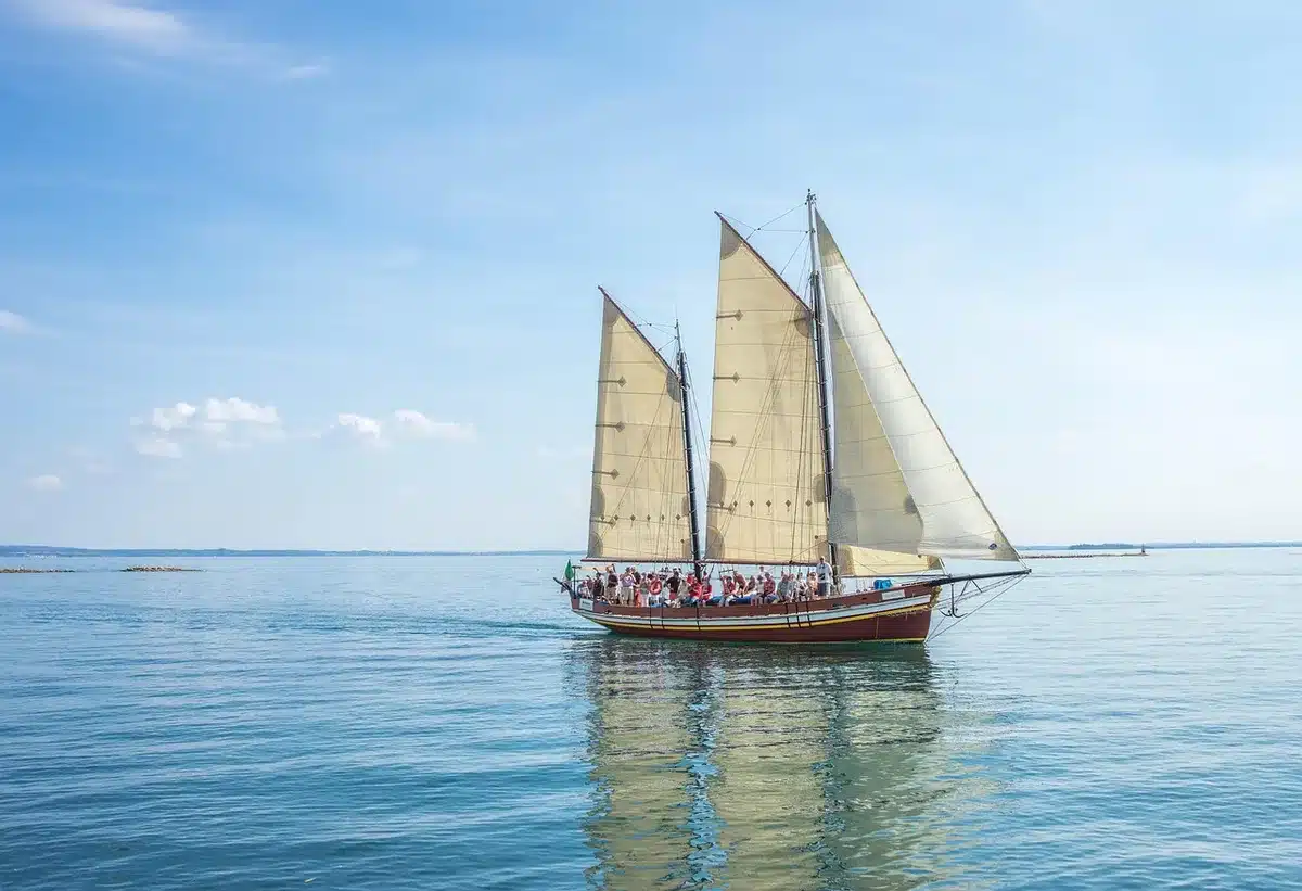 Ufer mit Sandstrand, Grasbüscheln, leerer Bank und Segelbooten auf einem großen See